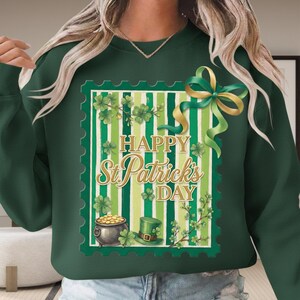 Puede incluir: Sudadera verde con un diseño del Día de San Patricio. El diseño presenta un sello postal con tréboles, una olla de oro, un sombrero verde y el texto "Happy St. Patrick's Day". Una cinta verde y dorada está en la parte superior derecha.