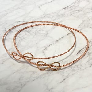 Pode incluir: Duas tiaras de arame cor de cobre com detalhes de nós infinitos. As tiaras são feitas de arame torcido e repousam sobre uma superfície de mármore. As tiaras são de uma cor metálica quente.