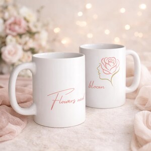 Könnte beinhalten: Zwei weiße Keramikbecher. Ein Becher hat den Text "Flowers need" in einer kursiven, korallenfarbenen Schrift. Der andere Becher hat ein Rosendesign mit dem Wort "bloom" in einer ähnlichen Korallenfarbe.