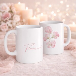 Könnte beinhalten: Zwei weiße Keramikbecher. Ein Becher hat den Text "Flowers need" in rosa Schrift. Der andere Becher zeigt ein rosa Blumendesign. Die Becher stehen auf einer rosa Oberfläche mit brennenden Kerzen im Hintergrund.