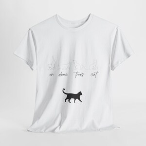 Può includere: T-shirt bianca con una silhouette nera di gatto e la scritta "un deux trois cat". Sopra il gatto, ci sono quattro disegni a tratto di gatti in diverse pose. Il testo è scritto a mano.