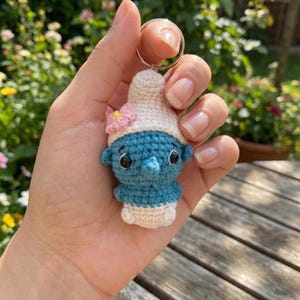May include: A handmade crochet keychain featuring a blue character with a white hat and a pink flower. The keychain is held by a hand, showcasing the detailed craftsmanship of the amigurumi design. The character has black eyes and a small nose.