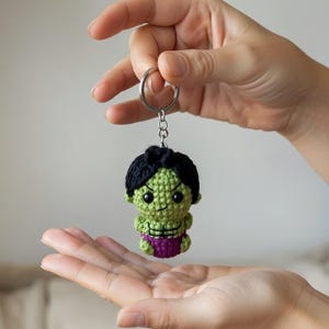 May include: A handmade crochet Hulk keychain. The Hulk figure is green with black hair and purple shorts. The keychain is attached to a silver metal ring.