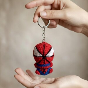 Op de afbeelding: Een handgemaakte Spider-Man sleutelhanger van gehaakt garen. De sleutelhanger heeft een rode kop met witte ogen en zwart web, een rood en blauw lichaam en een blauwe broek. Bevestigd aan een zilveren metalen ring en ketting.