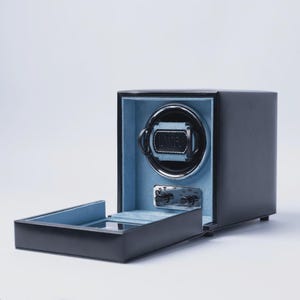 May include: A black watch winder with a light blue interior. The winder has a rectangular shape with a hinged door and a circular watch holder. The watch holder has the word "WOLF" on it.
