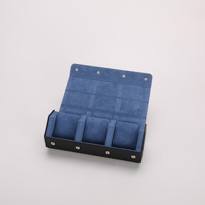 May include: Black leather watch case with a blue velvet interior. The case is open, revealing four watch compartments. The lid is secured with silver-colored snaps. The interior features a soft, blue velvet lining to protect the watches.
