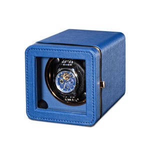 May include: A blue leather watch winder with a clear window displaying a silver and blue wristwatch. The winder has a textured exterior and gold-coloured accents. The watch features a blue face and a silver strap.