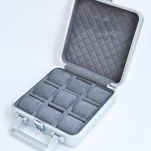 May include: Open silver-colored watch case with a gray quilted interior and nine watch pillows. The case has a handle and a latch closure. The interior is a soft gray fabric.