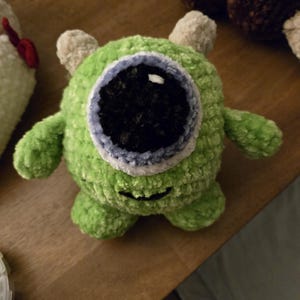 May include: A round, green, crocheted plush toy featuring a single large central eye with a black pupil and a light blue iris. The creature has two small beige horns on top, two short arms, two small legs, and a stitched black mouth.