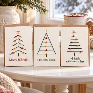 Può includere: Tre biglietti d'auguri color bianco sporco con disegni di alberi di Natale ricamati e piccole perline colorate. I biglietti riportano le scritte Merry & Bright, Joy to the World e A little Christmas cheer in inchiostro rosso e verde.