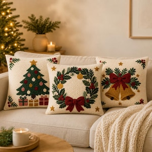 May include: Three square scatter cushions with cream-coloured textured fabric. The left cushion features a green Christmas tree with ornaments and gifts. The centre cushion shows a green wreath with a red bow. The right cushion displays two gold bells.
