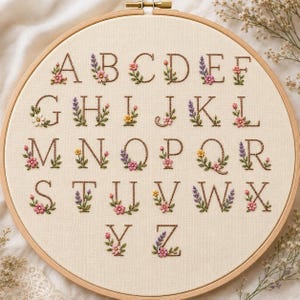 May include: A circular wooden embroidery hoop containing off-white fabric with the English alphabet embroidered in brown thread. Each letter is decorated with small, colorful floral motifs including pink, yellow, and purple flowers with green leaves.