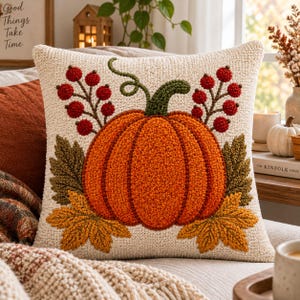 Könnte beinhalten: Ein quadratisches Zierkissen mit einer cremefarbenen, strukturierten Basis und einem zentralen Kürbismotiv in Orange. Der Kürbis ist mit grünen Stielen, braunen und gelben Herbstblättern sowie zwei Zweigen mit roten Beeren verziert.
