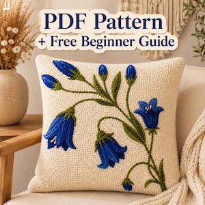 May include: A square cream-colored throw pillow featuring a textured punch needle design of blue bell-shaped flowers with green stems and leaves. The text PDF Pattern + Free Beginner Guide is displayed in dark blue letters above the pillow.