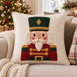 Könnte beinhalten: Ein quadratisches, cremefarbenes Zierkissen mit einem texturierten Punch-Needle-Design eines Nussknackers. Der Nussknacker hat einen dunkelgrünen Hut, eine rote Jacke mit goldenen Akzenten, einen weißen Bart und rosige Wangen.