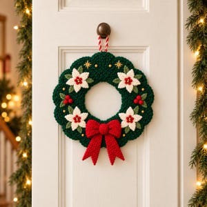 Könnte beinhalten: Ein runder, dunkelgrüner gehäkelter Kranz mit drei weißen Weihnachtssternen mit roten Zentren, roten Beerenclustern und kleinen goldenen Perlen. Eine große, strukturierte rote Schleife ist unten befestigt und wird an einer rot-weiß gestreiften Kordel aufgehängt.
