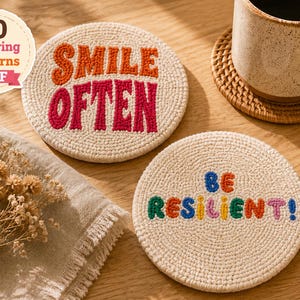 May include: Two round, off-white punch needle coasters featuring embroidered text. One coaster displays the orange and pink text "SMILE OFTEN," while the other features multicoloured "BE RESILIENT!" text. A circular badge indicates "20 Inspiring Patterns PDF."