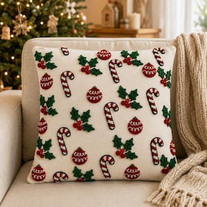 May include: A square throw pillow with an off-white fabric base featuring a repeating embroidered pattern of red and white candy canes, red Christmas ornaments with white swirl details, and green holly leaves with clusters of red berries.