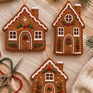 Könnte beinhalten: Drei braune Lebkuchenhaus-Anhänger aus Filz mit weißer Stickerei, grünem Blattwerk und roten Beeren. Jedes Haus verfügt über eine Aufhängeschlaufe, einen Schornstein, Fenster und eine Tür mit detaillierten, handgestickten Mustern.