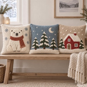 Könnte beinhalten: Drei quadratische Zierkissen mit texturierten Punch-Needle-Motiven. Das linke Kissen zeigt einen weißen Eisbären mit rotem Schal und blauen Schneeflocken. Das mittlere Kissen stellt eine blaue Nachtszene mit Tannen und einer Mondsichel dar. Das rechte Kissen zeigt ein rotes Haus mit schneebedecktem Dach.