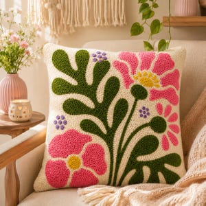 Könnte beinhalten: Ein quadratisches Zierkissen mit cremefarbener Basis und texturiertem Punch-Needle-Design. Das Muster zeigt große rosa Blumen mit gelben Mitten, dunkelgrüne blättrige Stängel und kleine Gruppen violetter Punkte auf der Stoffoberfläche.