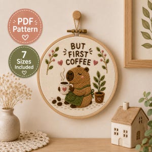 Könnte beinhalten: Ein runder Stickrahmen mit cremefarbenem Stoff, auf dem ein Capybara mit grüner Schürze und einer Kaffeetasse gestickt ist. Der Text lautet BUT FIRST COFFEE. Das Design enthält kleine rosa Herzen, Kaffeebohnen und eine Topfpflanze.
