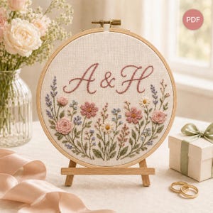 Könnte beinhalten: Ein runder Holzstickrahmen mit cremefarbenem Stoff, auf dem die Buchstaben A & H in altrosa Schreibschrift gestickt sind. Unter den Buchstaben befindet sich ein Blumenmuster aus rosa Rosen, kleinen blauen Blüten und grünem Blattwerk.