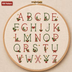 Könnte beinhalten: Ein runder hölzerner Stickrahmen mit cremefarbenem Stoff, auf dem ein Alphabet handgestickt ist. Jeder Buchstabe ist mit weihnachtlichen Motiven in Rot und Grün verziert, darunter Stechpalmen, Beeren, Schleifen, Schneeflocken und goldene Sterne.
