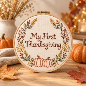 Könnte beinhalten: Ein runder Holzstickrahmen mit weißem Stoff, auf dem der Text My First Thanksgiving in Braun gestickt ist. Der Text ist von einem kreisförmigen Kranz aus gestickten Herbstblättern, Weizenähren und kleinen roten Beeren sowie zwei Kürbissen umgeben.