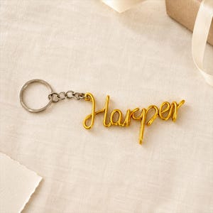 May include: A gold-colored metal keychain featuring the name Harper written in a continuous cursive wire script. The name is attached to a standard circular silver-toned metal split ring by a short chain of three small, interconnected metal links.