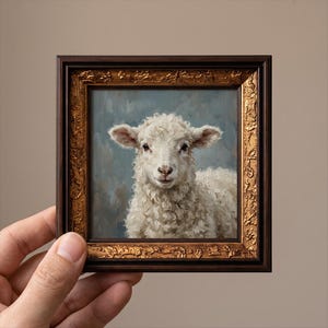 Könnte beinhalten: Ein kleines quadratisches Ölgemälde eines weißen Lamms mit lockigem Fell vor einem gedämpften blaugrauen Hintergrund. Das Kunstwerk ist in einem verzierten, dunkelbraunen und goldfarbenen Rahmen mit komplexen Prägungen gefasst.
