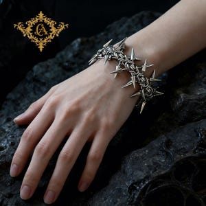 May include: A silver bracelet with a thorny design. The bracelet has multiple sharp spikes and is worn on a wrist. The bracelet is made of metal and has a gothic or punk aesthetic.