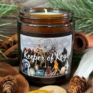 May include: A brown glass jar candle with a white label that reads "Keeper of Keys." The label features illustrations of keys, a dog, and a campfire. The candle is surrounded by pinecones, feathers, and wood slices.