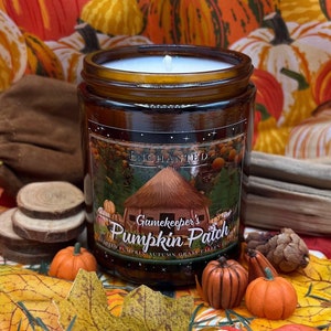 Könnte beinhalten: Eine braune Glas-Kerze mit einem weißen Docht, beschriftet mit "Gamekeeper's Pumpkin Patch". Das Etikett zeigt eine kleine Hüttenillustration. Die Kerze ist umgeben von Miniaturkürbissen, Holzscheiben und Herbstblättern, mit einem Kürbis-Themenhintergrund.