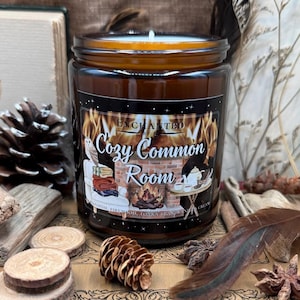 May include: A brown glass candle jar with a black label that reads "Enchanted by Cozy Common Room" with a graphic of a fireplace, an owl, and a cup of tea. The candle is surrounded by pine cones, a book, and a feather.