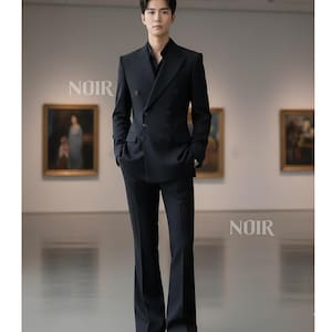 以下が含まれることがあります： 黒のダブルブレストスーツ、フレアパンツ付き。ノッチドラペルと複数のボタンが付いています。「NOIR」の文字が壁に白で印刷されています。人物は絵画の前に立っています。