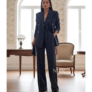 May include: A navy blue pinstripe suit, consisting of a double-breasted blazer with gold buttons and wide-leg trousers. The suit is worn with a black clutch. The outfit is suitable for business or formal occasions.
