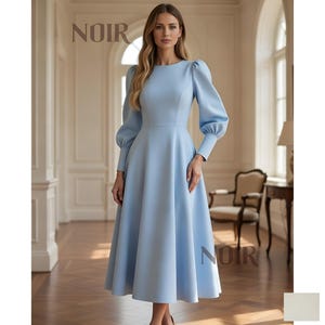 May include: A pale blue dress with long sleeves and a flared skirt. The dress has a round neckline and a fitted bodice. The sleeves have a puffed design at the shoulders. The word "NOIR" is printed in a serif font at the top and bottom of the image.