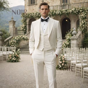 May include: A formal ivory suit with a tailcoat, vest, and trousers. The suit is paired with a black bow tie and a white dress shirt. The word "NOIR" is displayed in the upper left corner. The background features a stone building and floral arrangements.
