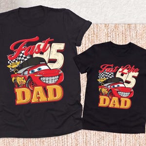 May include: Two black short-sleeve t-shirts featuring a graphic of a red race car wearing a black baseball cap and braces. The design includes a checkered flag, the number 95, and the text Fast DAD on the larger shirt and Fast Like DAD on the smaller.