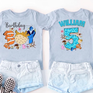 May include: Two light blue t-shirts with ocean-themed graphics. The left shirt features the text "Birthday BOY" with fish illustrations. The right shirt displays the name "WILLIAM" above a large number "5" filled with a blue water pattern.