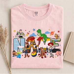 K&ouml;nnte beinhalten: Ein hellrosa T-Shirt mit einem Grafikdruck von Toy Story-Figuren wie Buzz Lightyear, Woody, Jessie, Rex und Slinky Dog, die um das Wort NAME in gro&szlig;en Blockbuchstaben angeordnet sind, mit einem Comfort Colors Markenetikett am Halsausschnitt.