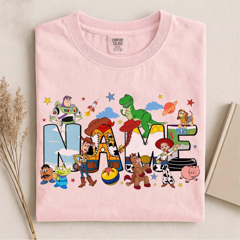 K&ouml;nnte beinhalten: Ein hellrosa T-Shirt mit einem Grafikdruck von Toy Story-Figuren wie Buzz Lightyear, Woody, Jessie, Rex und Slinky Dog, die um das Wort NAME in gro&szlig;en Blockbuchstaben angeordnet sind, mit einem Comfort Colors Markenetikett am Halsausschnitt.