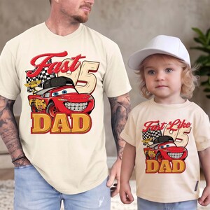 May include: Two cream-colored t-shirts featuring a red race car graphic with a black baseball cap, a checkered flag, and the number 95. The adult shirt displays the text Fast DAD, while the child shirt displays the text Fast Like DAD.