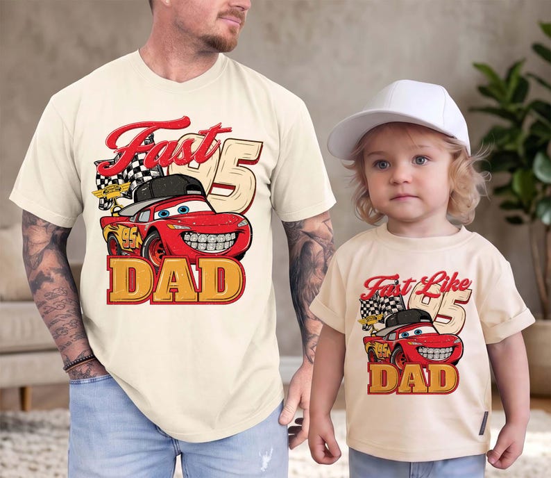 May include: Two cream-colored t-shirts featuring a red race car graphic with a black baseball cap, a checkered flag, and the number 95. The adult shirt displays the text Fast DAD, while the child shirt displays the text Fast Like DAD.