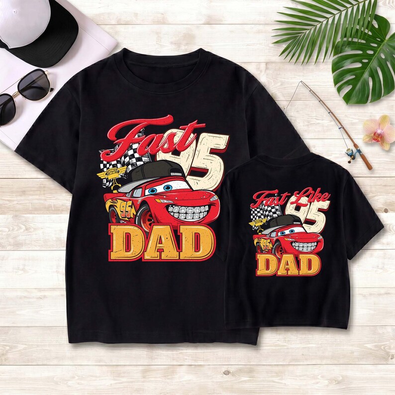 May include: Two black t-shirts featuring a red race car graphic wearing a black baseball cap and braces. The larger shirt displays the text Fast 95 DAD, while the smaller shirt displays the text Fast Like 95 DAD, both with a checkered flag.