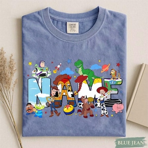 K&ouml;nnte beinhalten: Ein blaues T-Shirt mit einem Grafikdruck verschiedener animierter Figuren aus der Toy Story-Reihe. Das Wort NAME ist in gro&szlig;en, bunten Buchstaben dargestellt, die jeweils mit Mustern und Bildern der Figuren wie Woody, Buzz Lightyear, Jessie, Rex und Slinky Dog gef&uuml;llt sind.