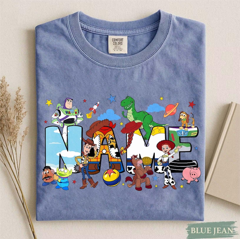 K&ouml;nnte beinhalten: Ein blaues T-Shirt mit einem Grafikdruck verschiedener animierter Figuren aus der Toy Story-Reihe. Das Wort NAME ist in gro&szlig;en, bunten Buchstaben dargestellt, die jeweils mit Mustern und Bildern der Figuren wie Woody, Buzz Lightyear, Jessie, Rex und Slinky Dog gef&uuml;llt sind.