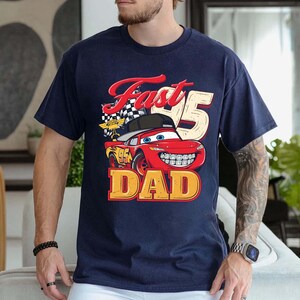 May include: A navy blue short-sleeve t-shirt featuring a graphic of a red race car with a black baseball cap, a checkered flag, and the text Fast 95 DAD. The car has blue eyes, a wide smile with visible teeth, and the number 95 on its side.