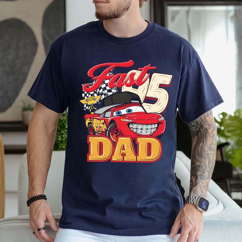 May include: A navy blue short-sleeve t-shirt featuring a graphic of a red race car with a black baseball cap, a checkered flag, and the text Fast 95 DAD. The car has blue eyes, a wide smile with visible teeth, and the number 95 on its side.